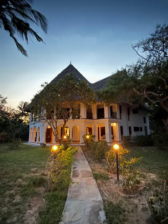 Enkare Bofa lit up at dusk, a lantern-lined stone path leading across the lawn to the arched veranda