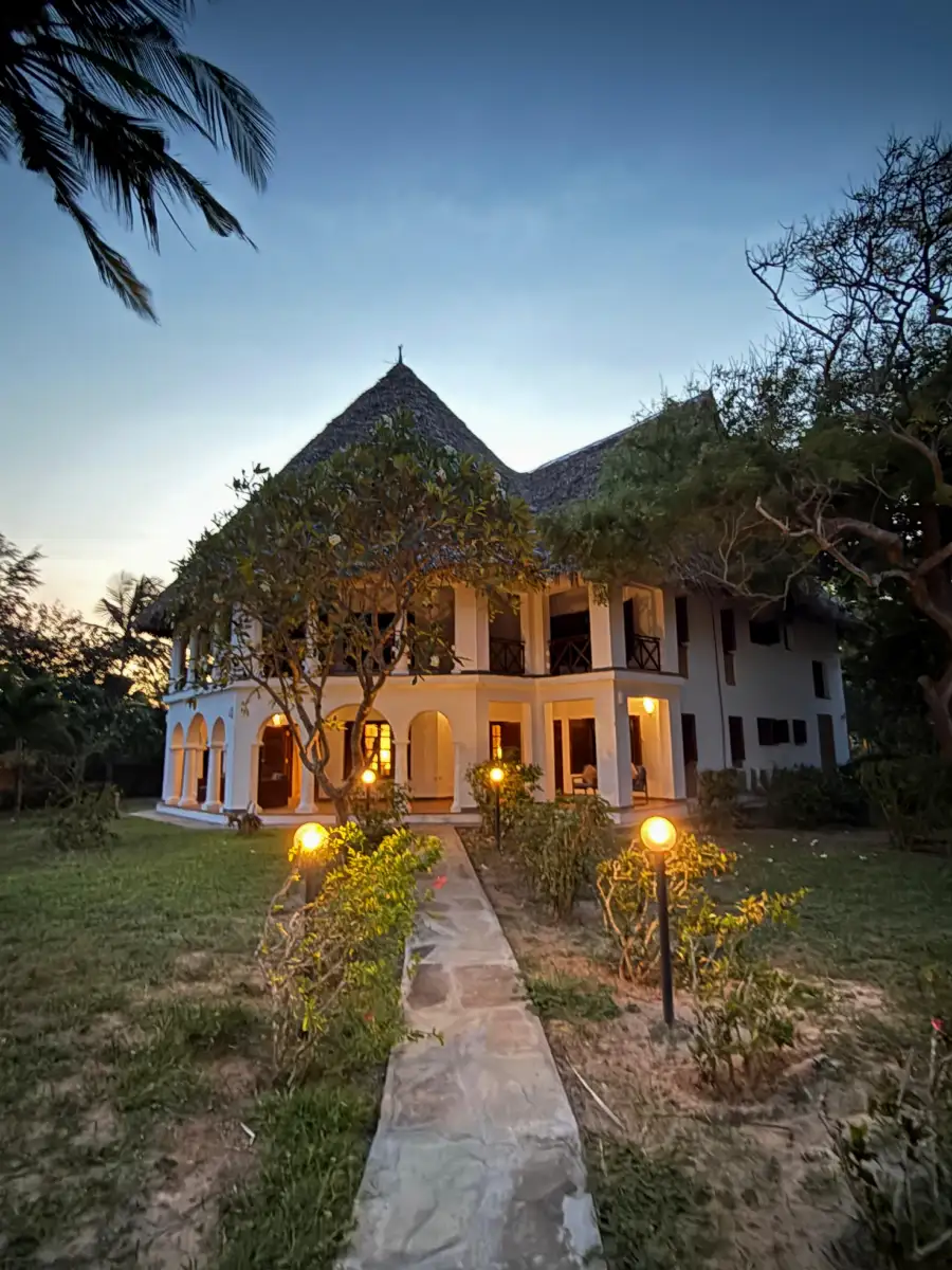 Enkare Bofa lit up at dusk, a lantern-lined stone path leading across the lawn to the arched veranda